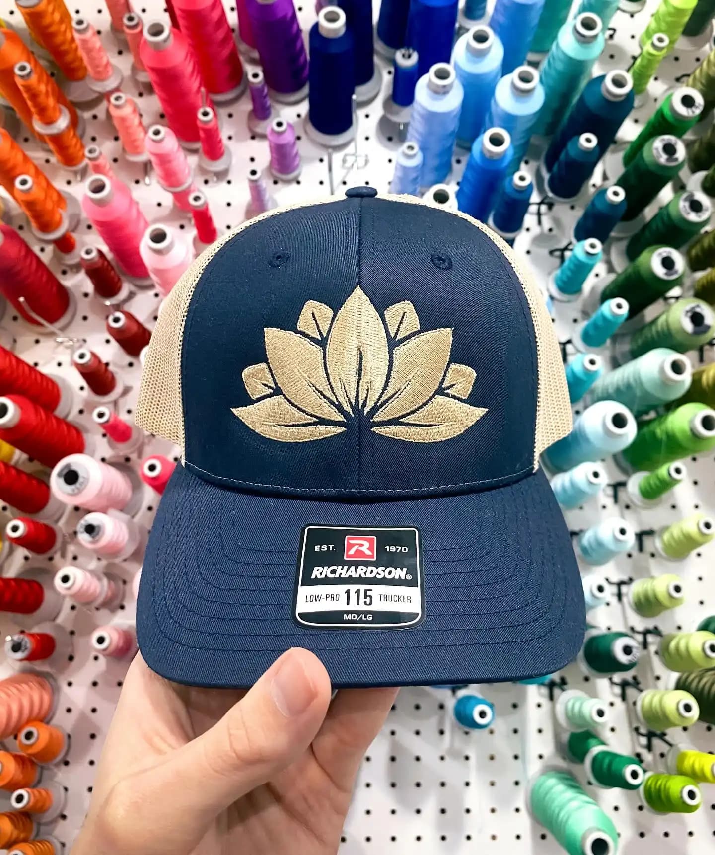 Gold lotus flower logo on a navy Richardson 115 Low-Pro Trucker hat, utilizing 100% manual digitizing for fine-detail boutique headwear.