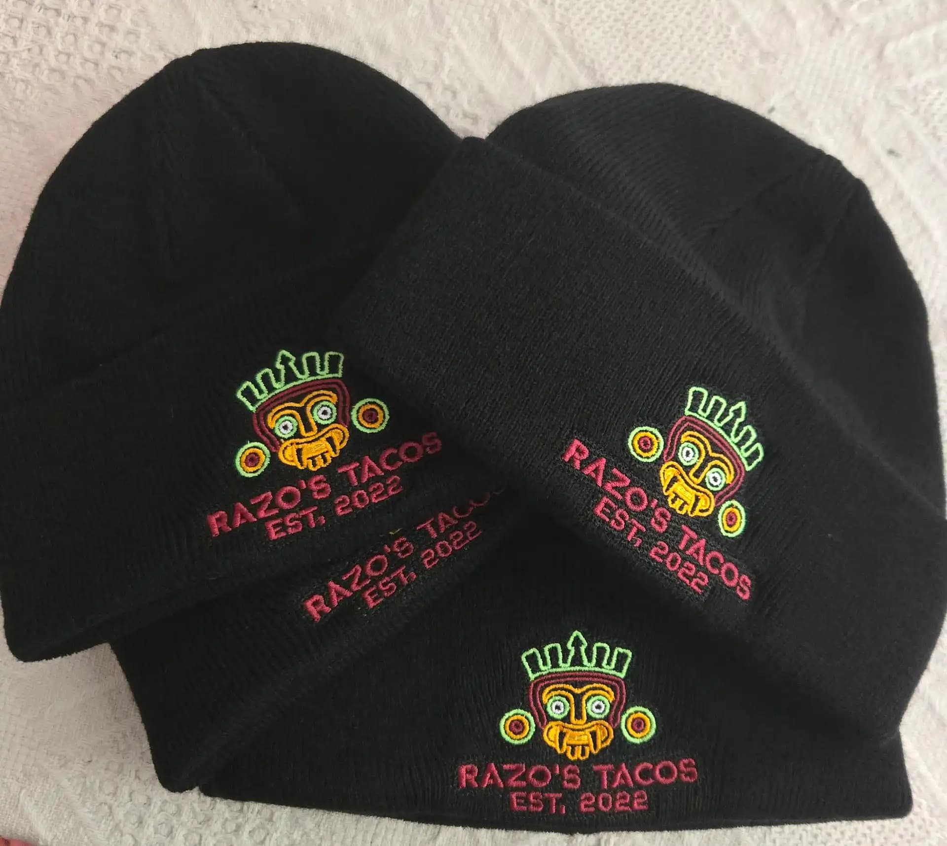 Razo’s Tacos mascot logo on black knit beanies with neon green and red thread, showcasing 100% manual digitizing for vibrant restaurant apparel.