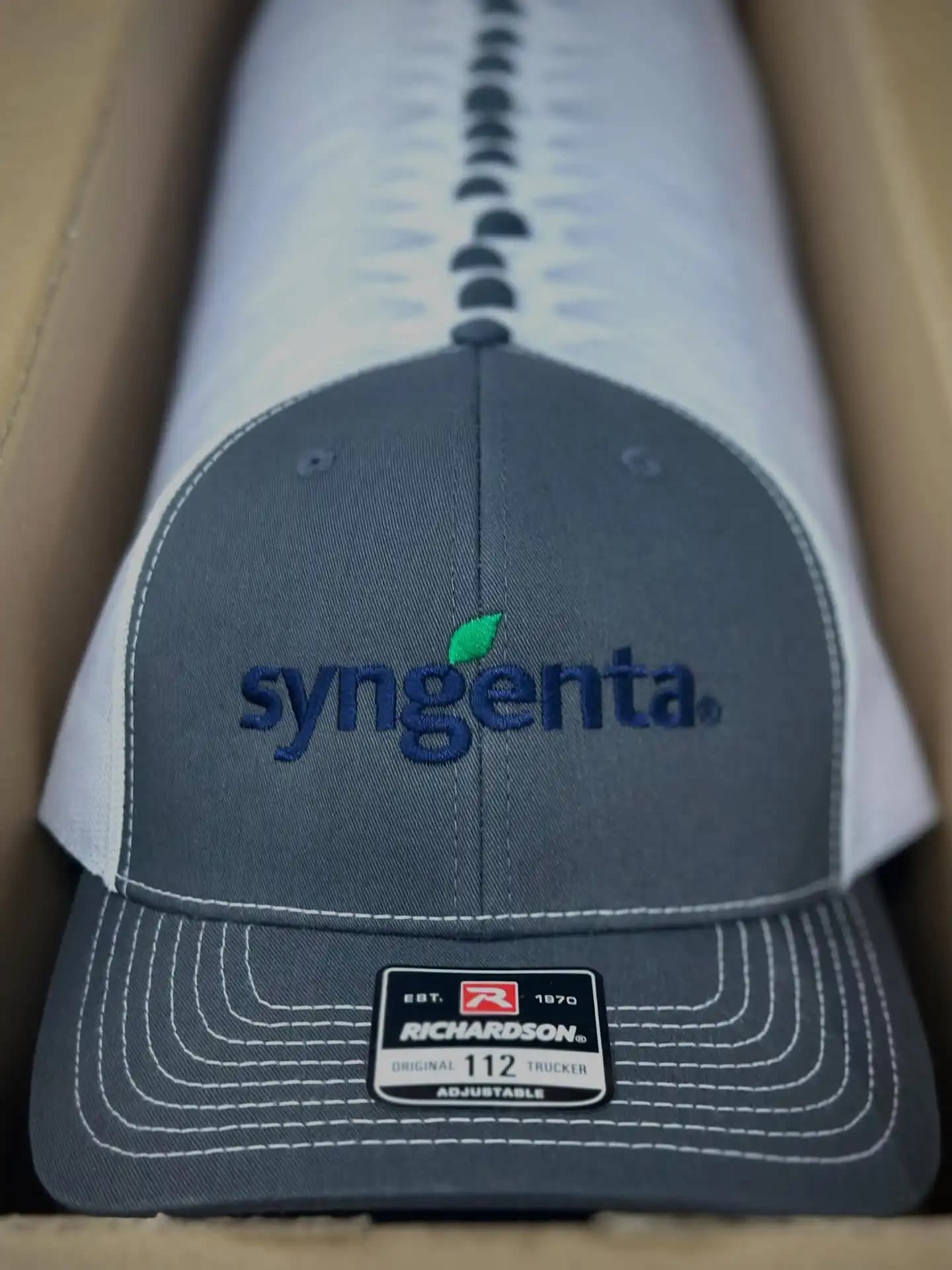 Syngenta corporate logo for bulk production on Richardson 112 trucker hats, utilizing 100% manual digitizing to ensure brand consistency across charcoal and white fabrics.